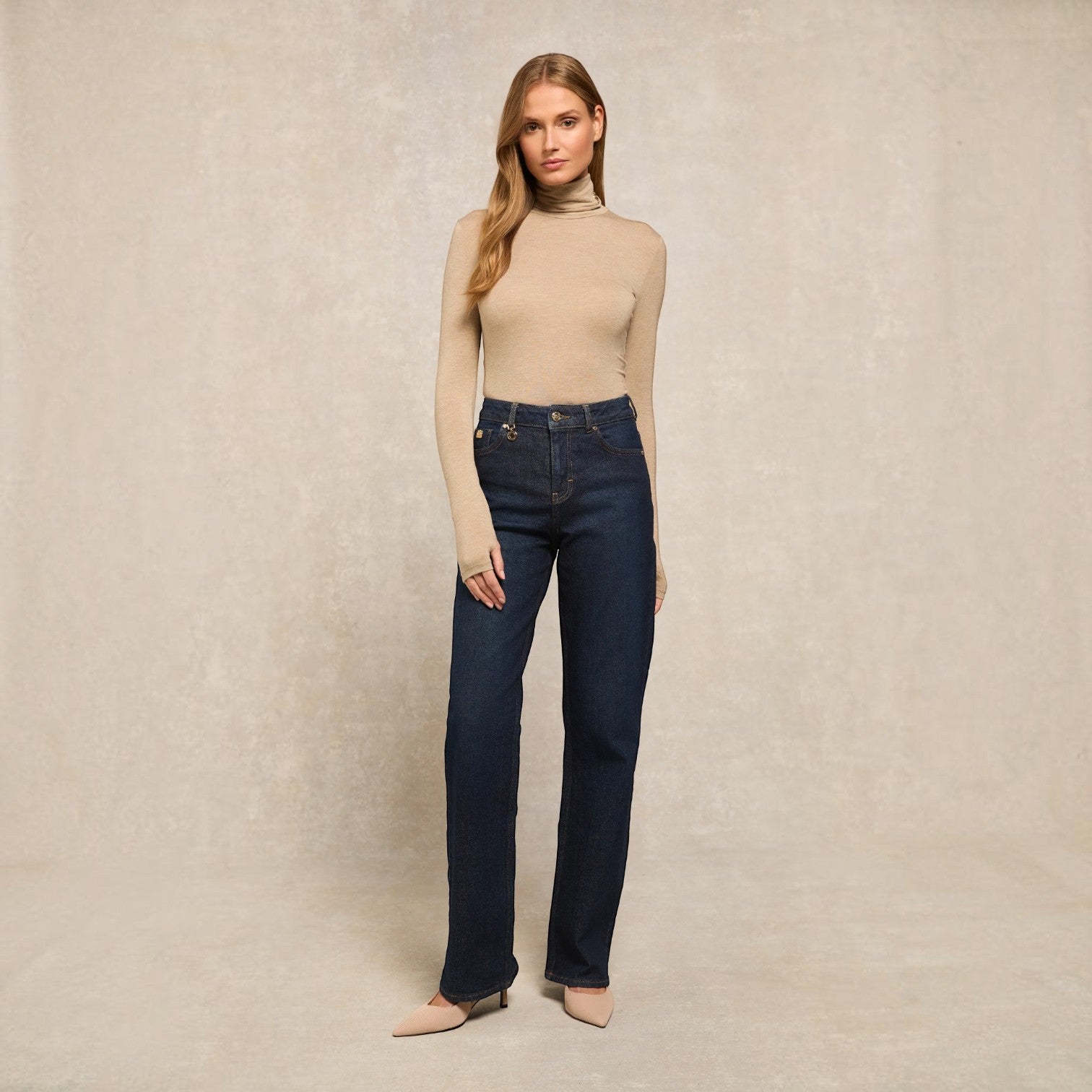 A woman stands against a textured beige studio backdrop wearing a beige turtleneck sweater, dark blue straight-leg jeans, and beige pointed-toe heels.