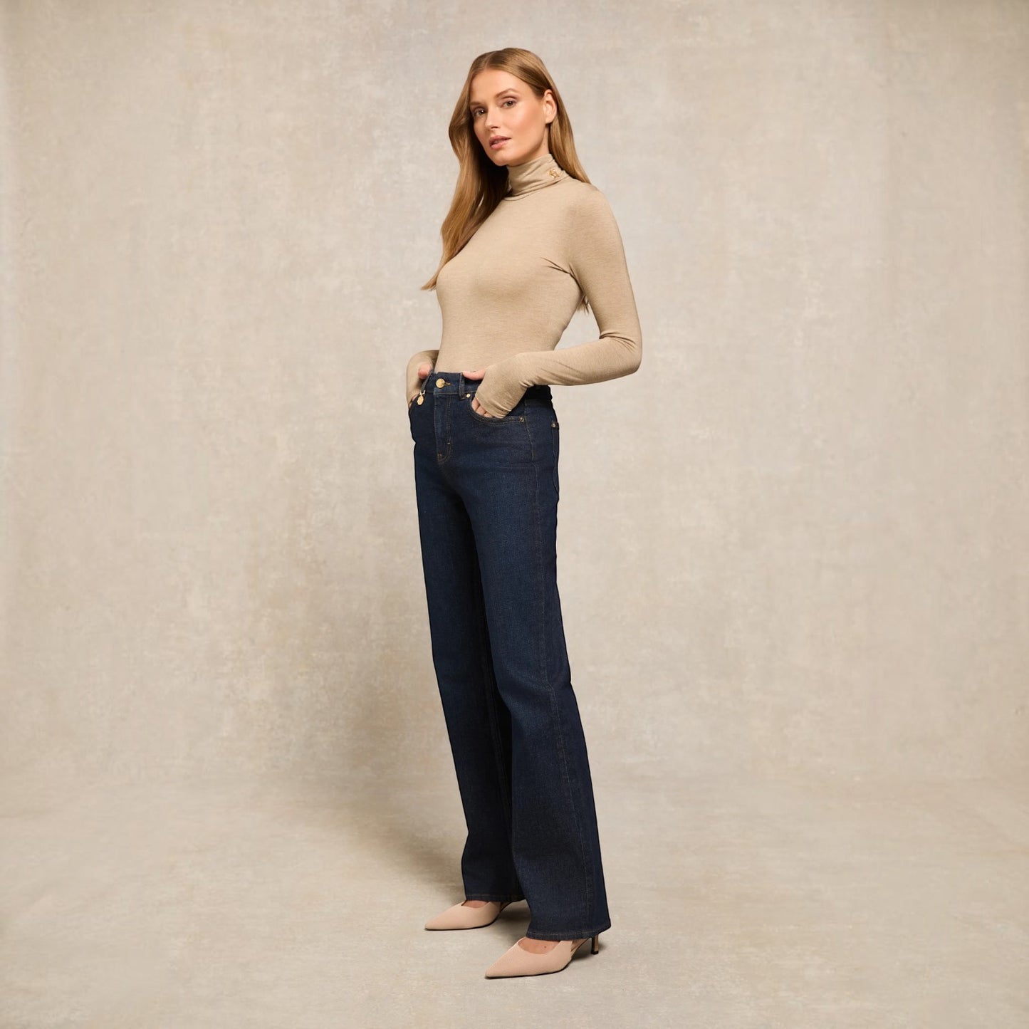 A woman modeling a beige long-sleeve turtleneck sweater and dark blue wide-leg jeans against a textured beige studio backdrop.