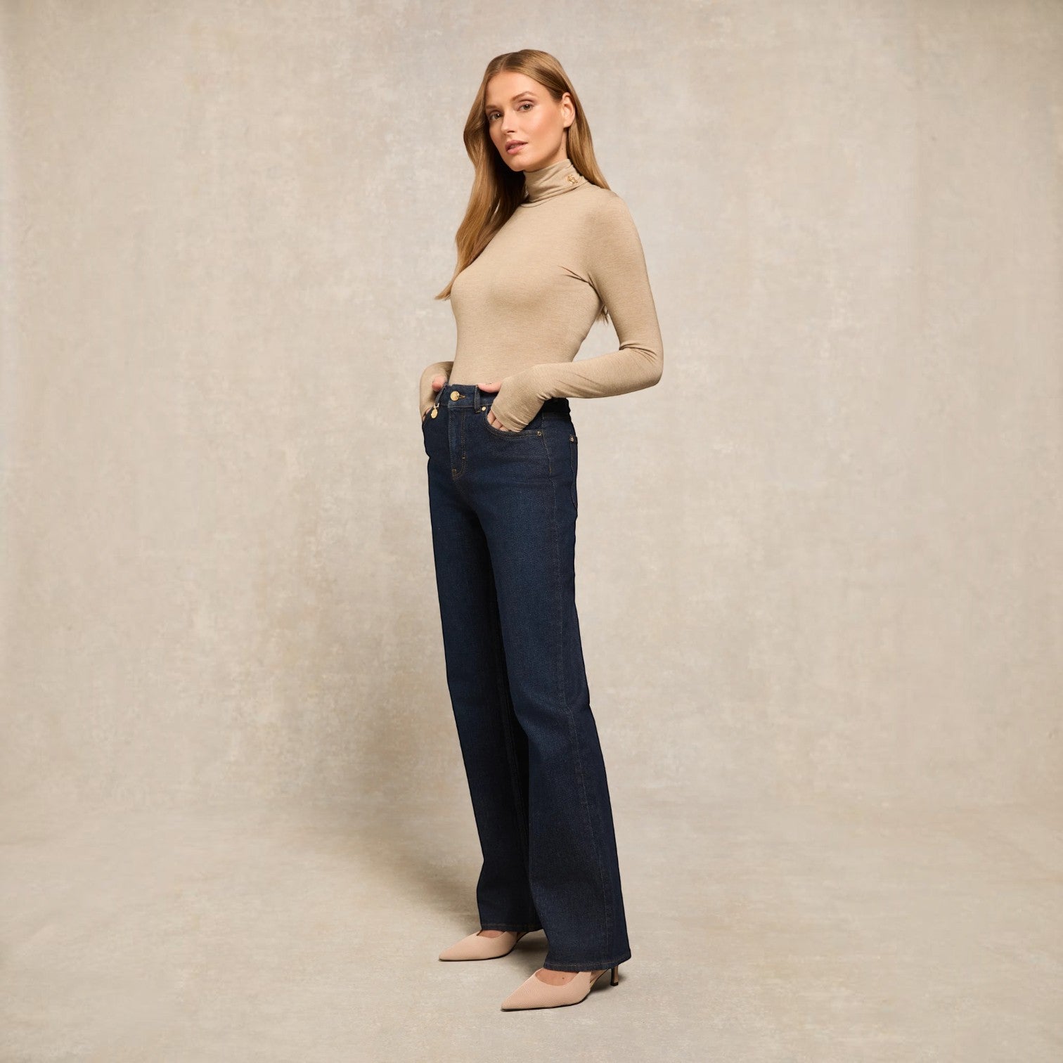 A woman modeling a beige long-sleeve turtleneck sweater and dark blue wide-leg jeans against a textured beige studio backdrop.