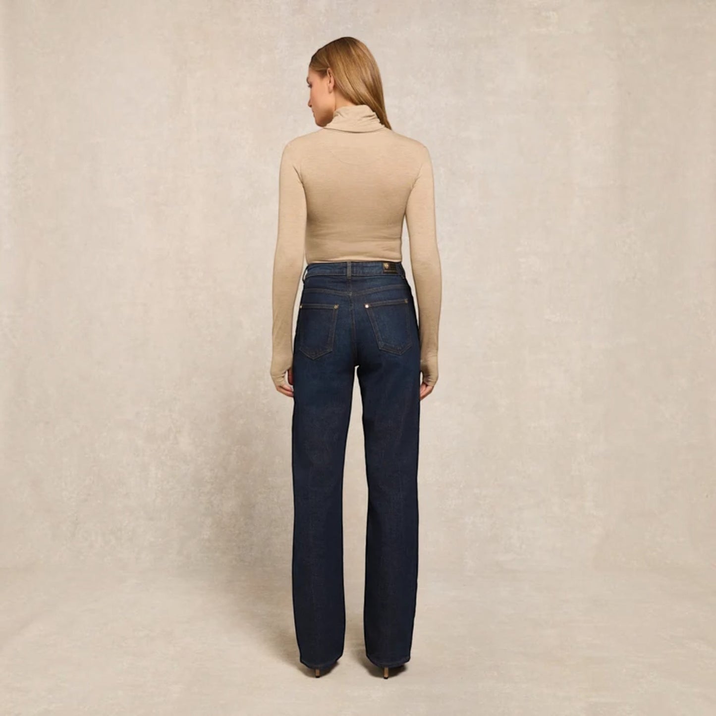 A woman stands with her back to the camera wearing a beige ribbed turtleneck top and dark blue straight-leg jeans against a textured beige studio backdrop.