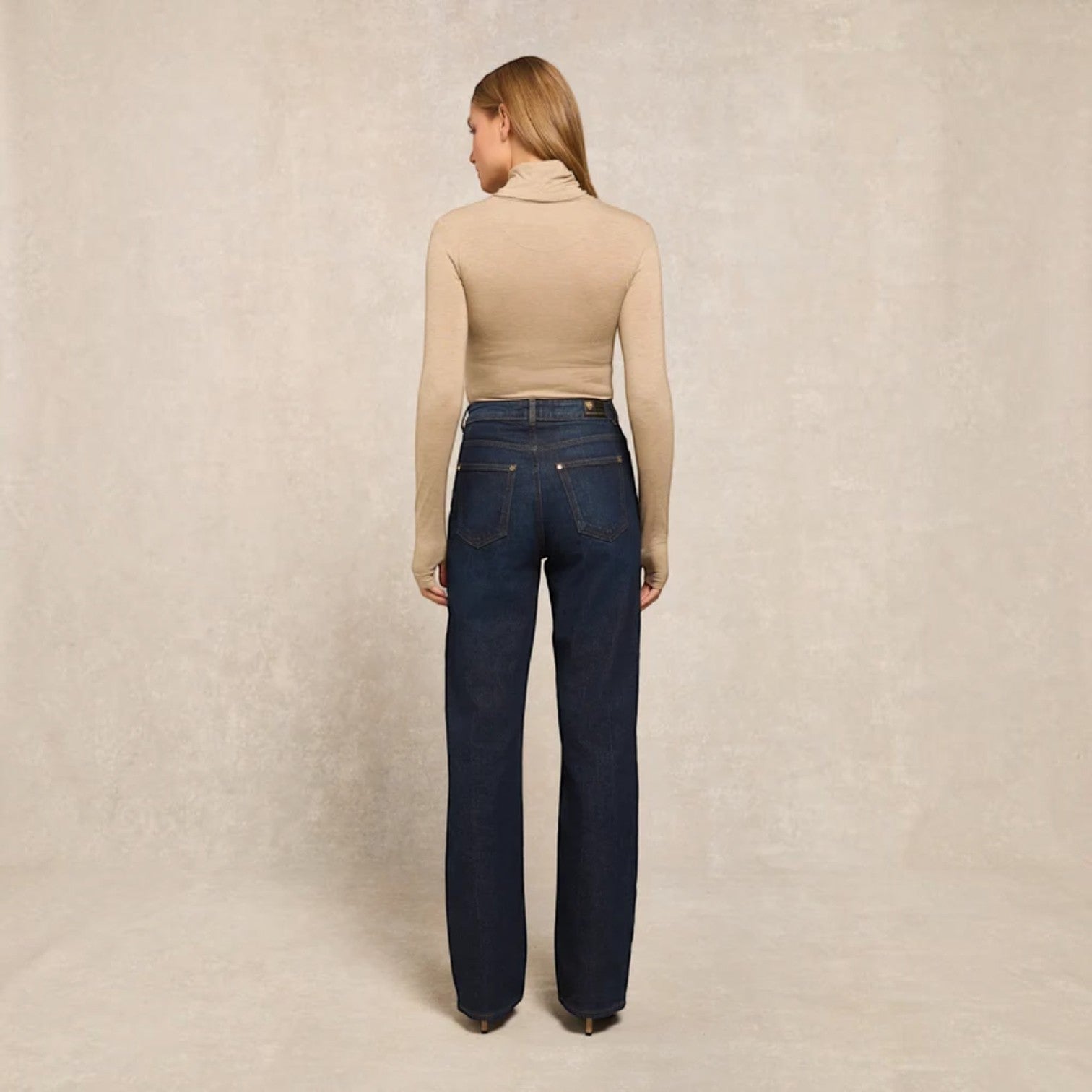 A woman stands with her back to the camera wearing a beige ribbed turtleneck top and dark blue straight-leg jeans against a textured beige studio backdrop.