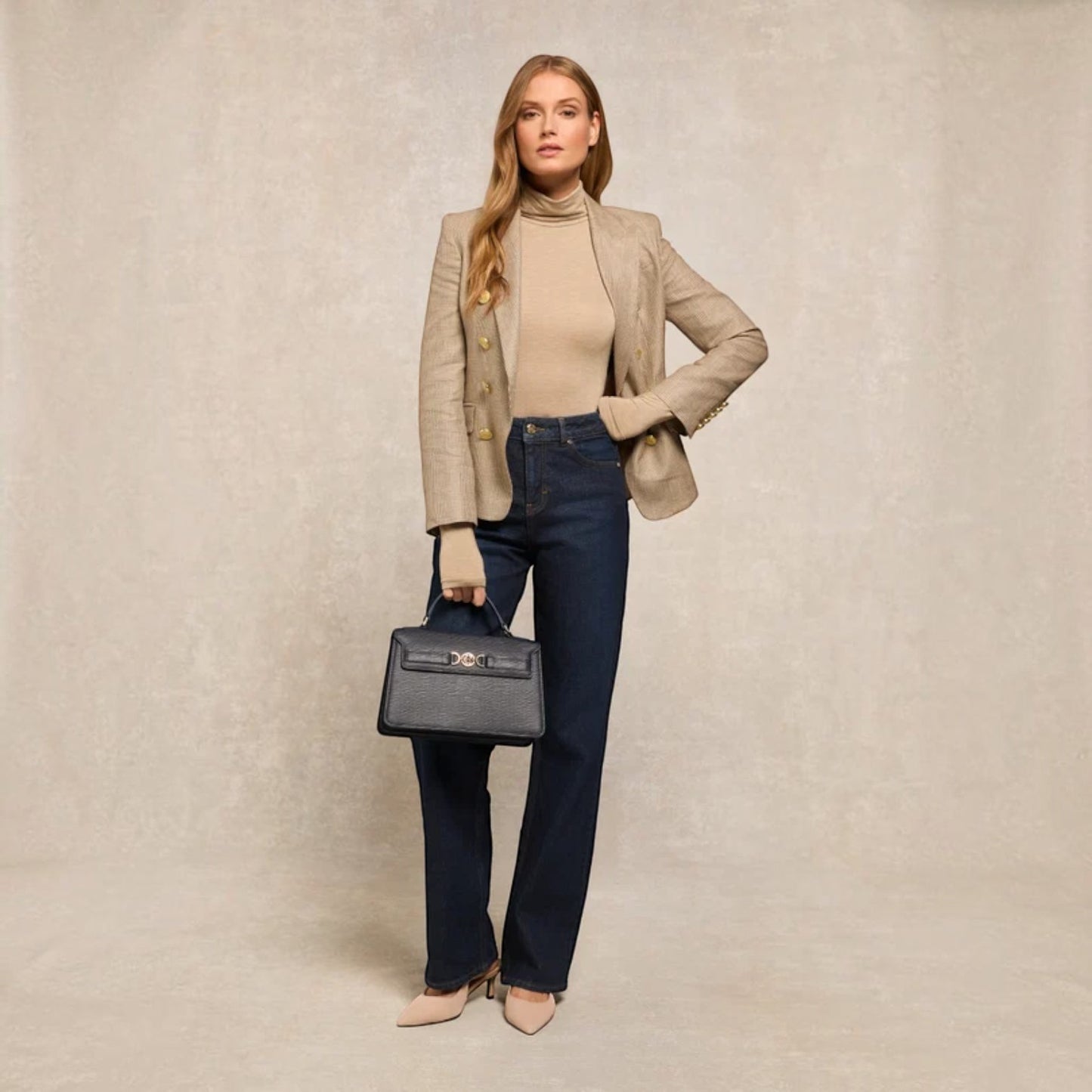A woman models a beige double-breasted blazer, beige turtleneck, dark blue straight-leg jeans, and a dark gray structured handbag against a textured beige studio backdrop.