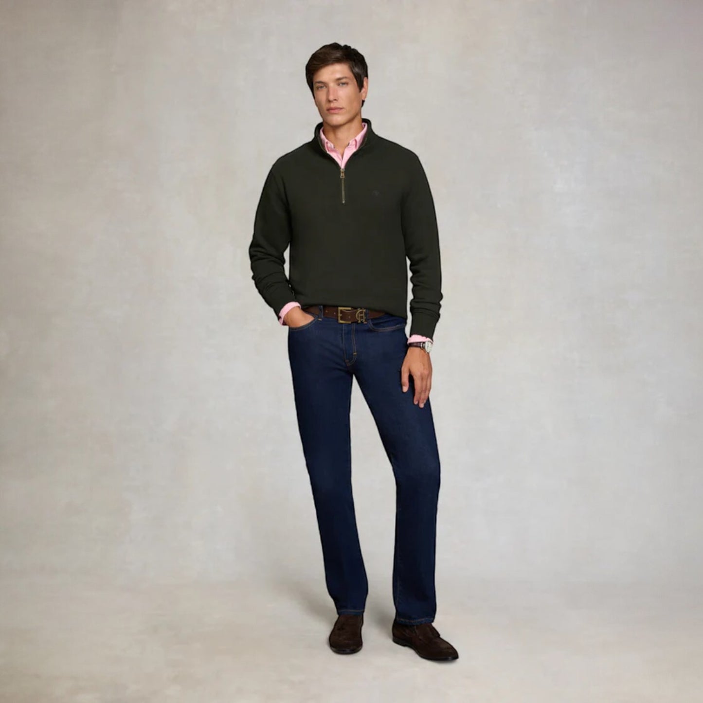 Man wearing a dark green sweater, pink shirt, blue jeans, and brown shoes on a plain background