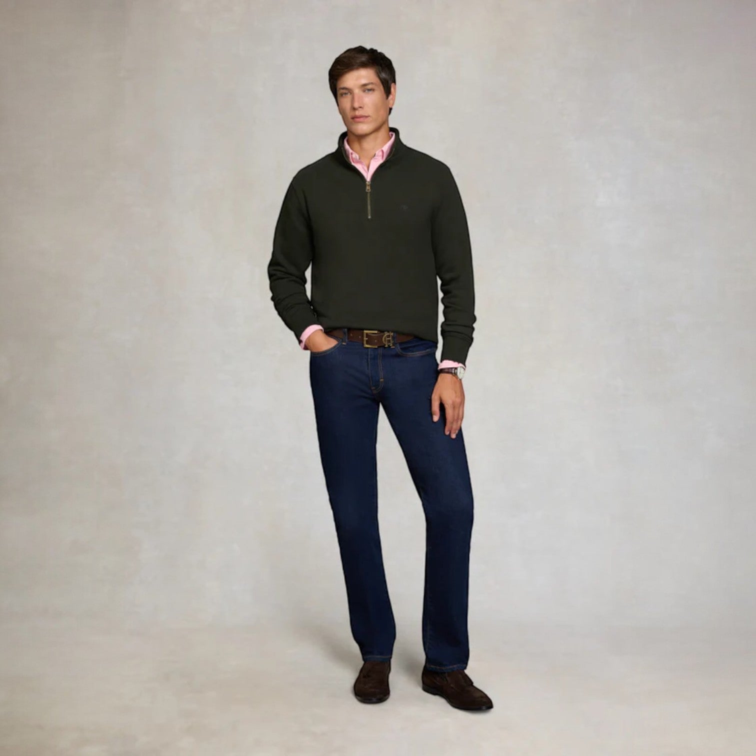 Man wearing a dark green sweater, pink shirt, blue jeans, and brown shoes on a plain background