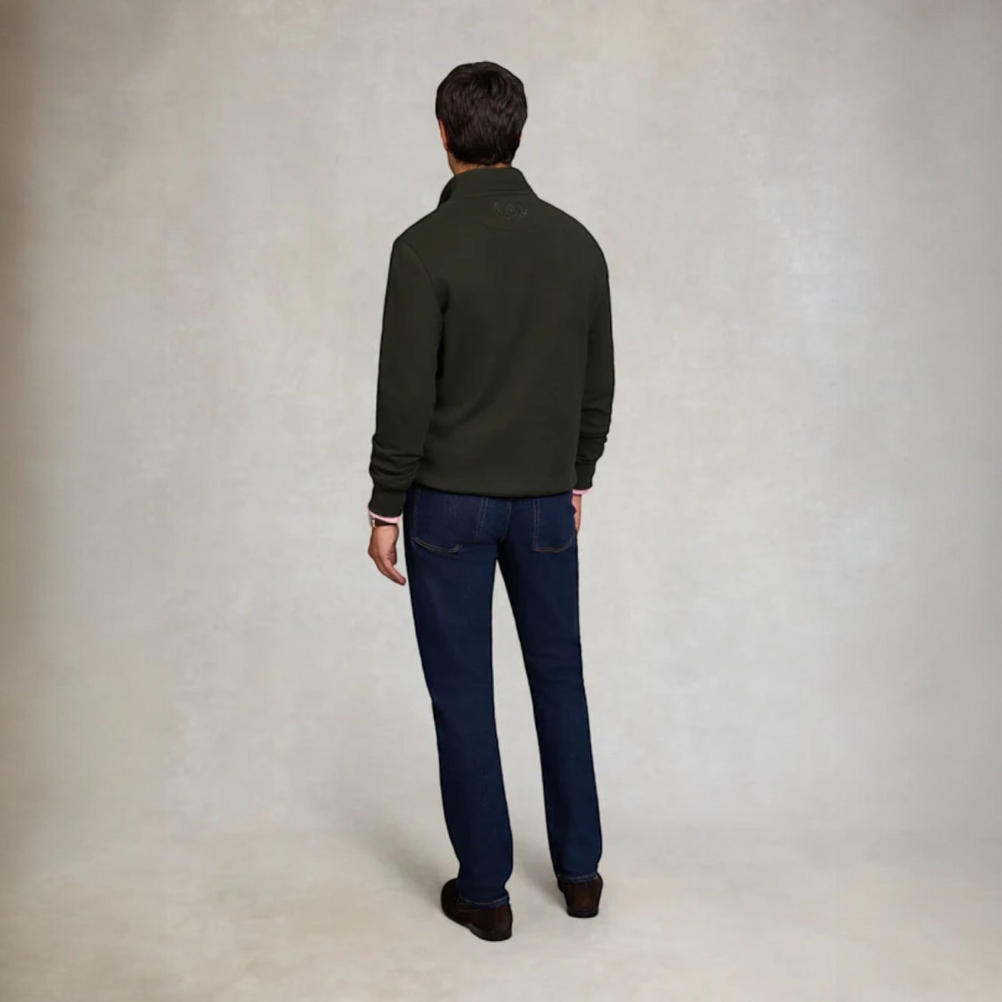 Man wearing a green jacket and blue jeans standing against a plain background