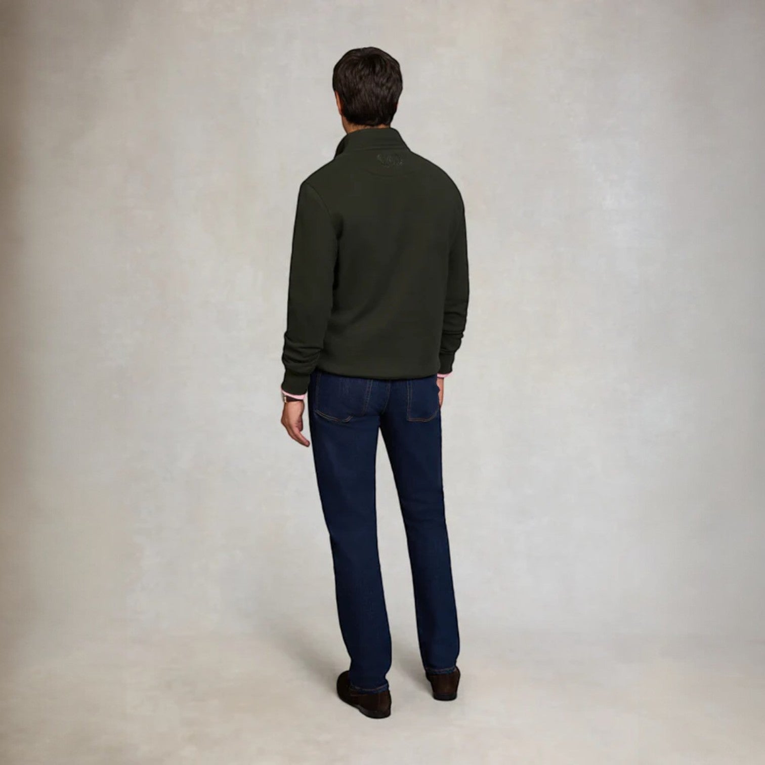 Man wearing a green jacket and blue jeans standing against a plain background