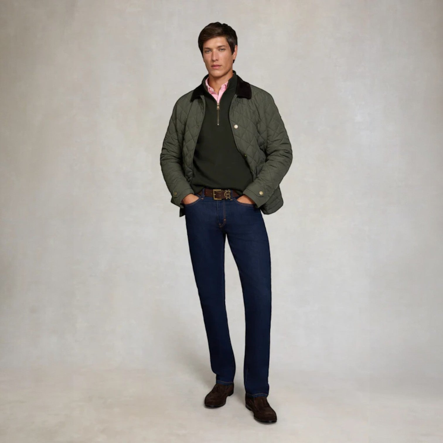 Man wearing a green jacket, dark sweater, blue jeans, and brown shoes on a plain background
