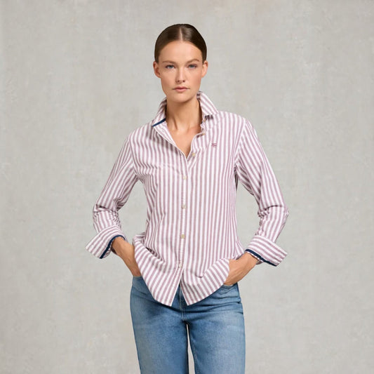 A woman modeling a long-sleeve button-up shirt with vertical pink and white stripes, paired with blue jeans, against a textured light gray background.