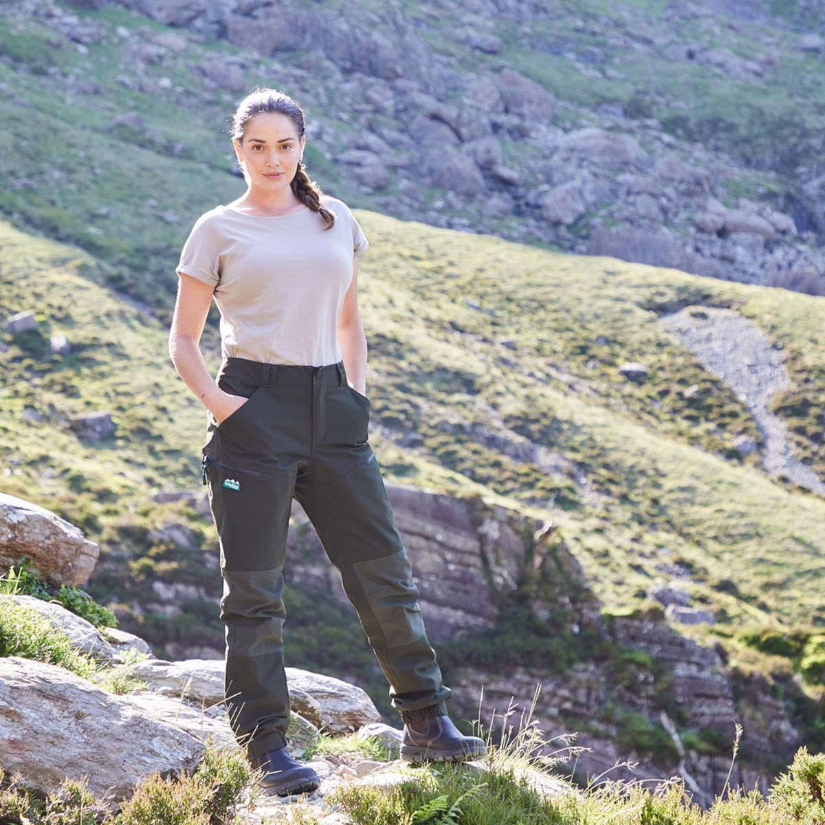 A woman stands outdoors on a rocky grassy hillside wearing dark olive green technical outdoor pants and a beige t-shirt.