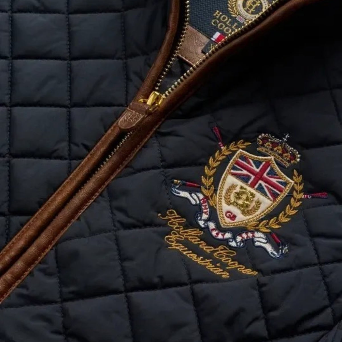 Close-up of a quilted navy jacket with a brown leather zipper trim and an embroidered crest on the chest. The crest includes a crown, shield with Union Jack, laurel wreath, and gold script reading 'Holland Cooper Equestrian'.