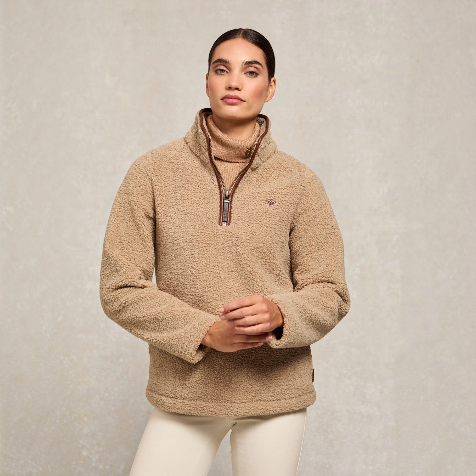 A woman wearing a beige sherpa fleece quarter-zip pullover sweater with a brown collar and zipper trim, paired with white pants, standing against a textured light gray studio background.