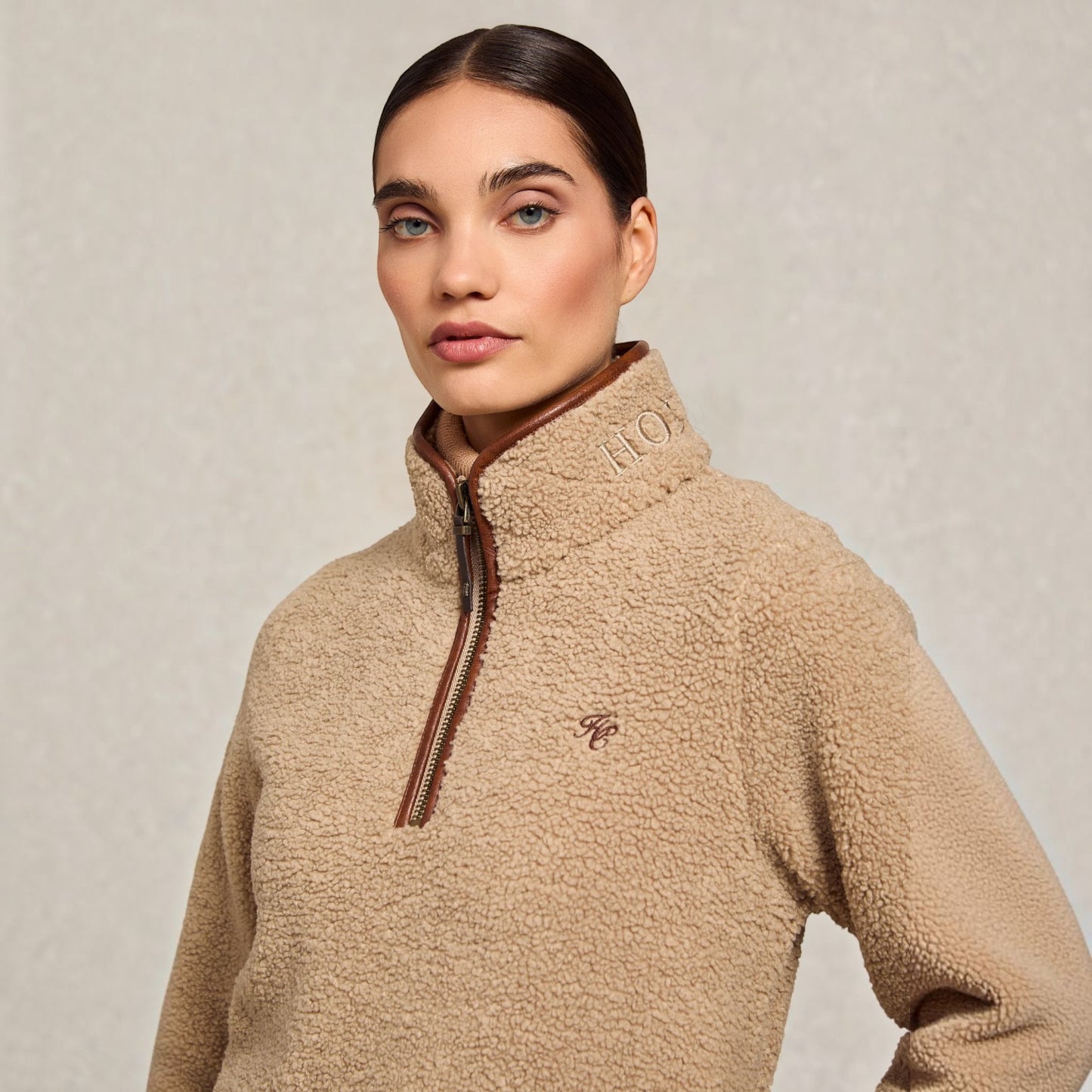 A woman wearing a beige sherpa fleece quarter-zip pullover sweater with brown trim and zipper details, photographed against a neutral textured light gray background.