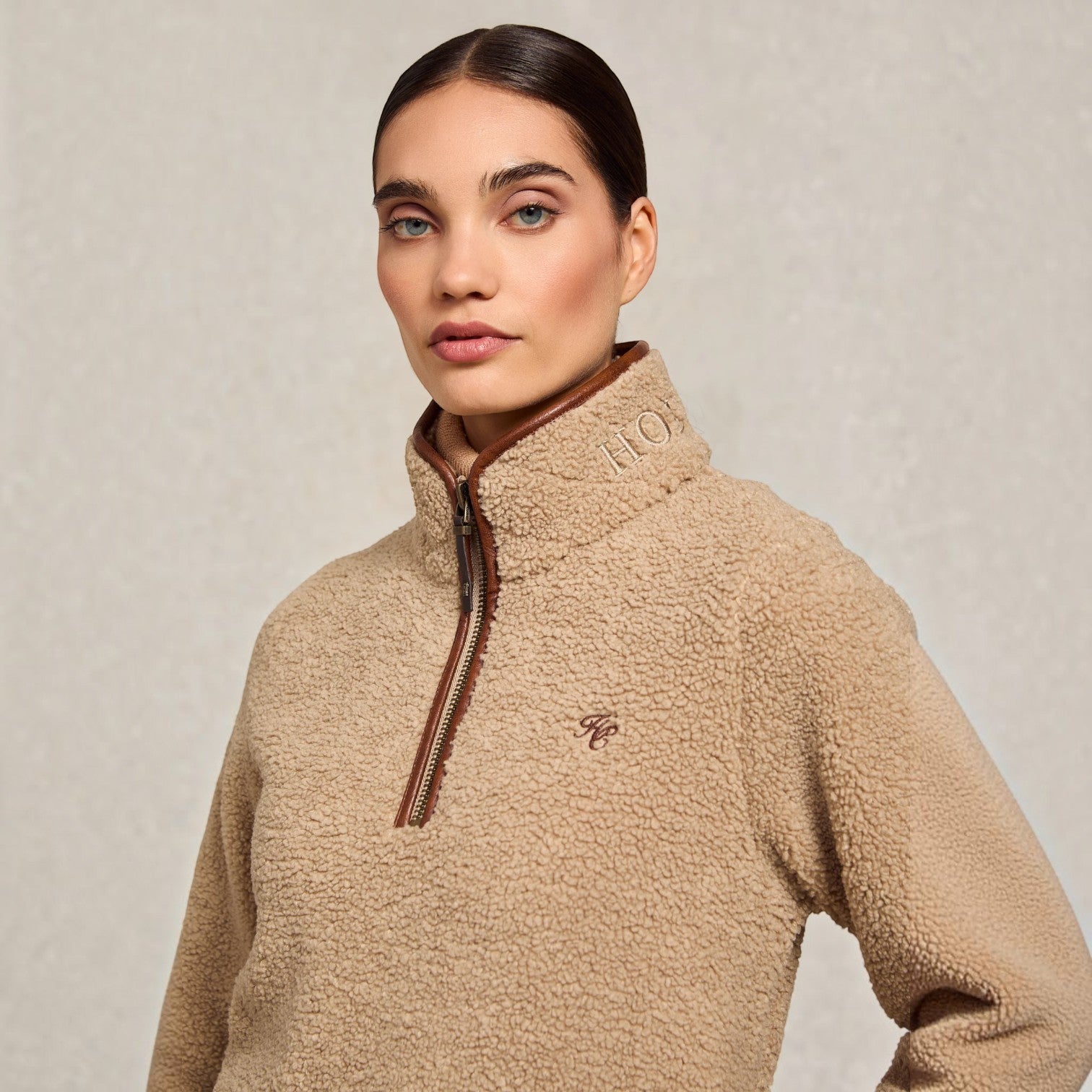 A woman wearing a beige sherpa fleece quarter-zip pullover sweater with brown trim and zipper details, photographed against a neutral textured light gray background.