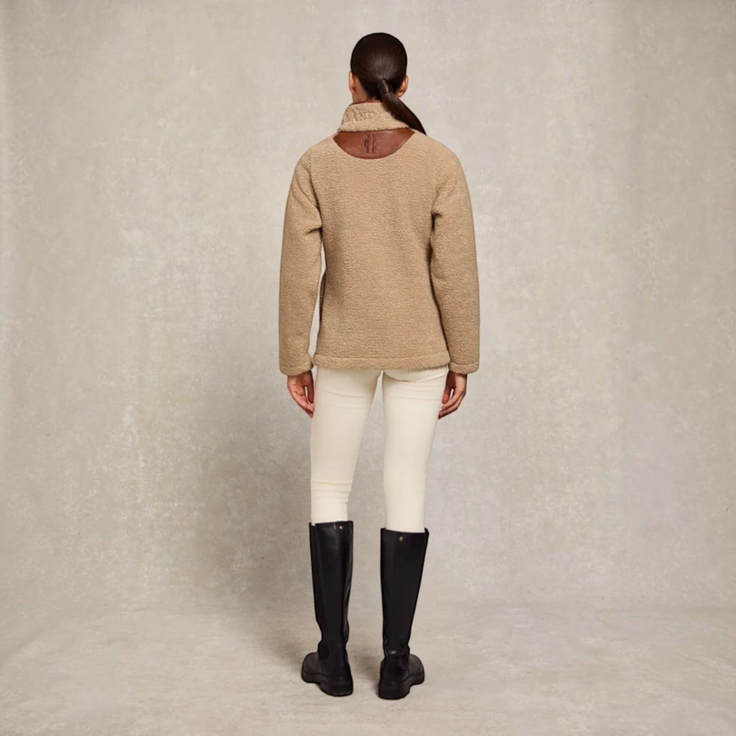 A model viewed from the back wearing a beige bouclé knit sweater with a brown leather patch at the collar, white riding breeches, and black riding boots, set against a textured light gray studio backdrop.