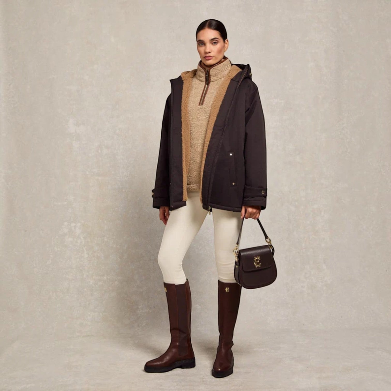 A model stands against a textured light gray studio backdrop wearing a dark brown hooded parka over a beige knit top, white pants, and dark brown knee-high boots, while carrying a dark brown handbag.