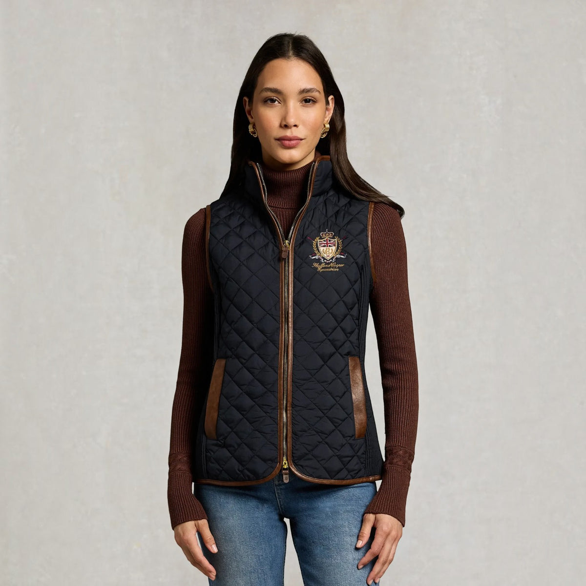 A woman modeling a navy quilted gilet vest with brown leather trim and a gold crest logo on the chest, worn over a brown ribbed turtleneck sweater and blue jeans against a textured light gray background.