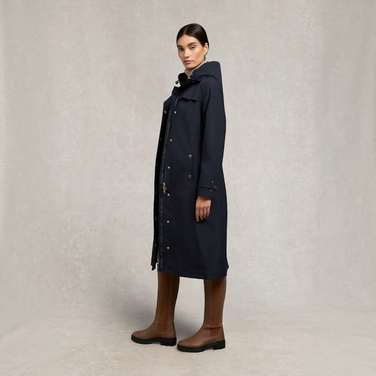 A model wearing a long navy blue hooded coat with gold buttons, paired with brown knee-high boots, standing against a textured light gray studio backdrop.