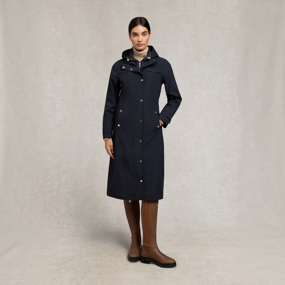 A woman modeling a long navy hooded coat with gold-tone buttons, paired with brown knee-high boots, against a neutral textured studio backdrop.