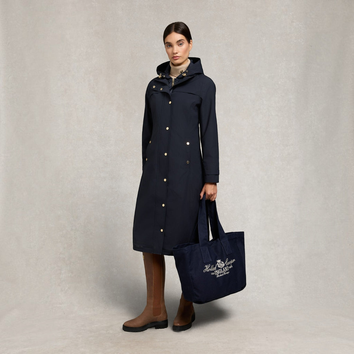 A woman models a long navy hooded coat with gold buttons, holding a matching navy tote bag with a white embroidered logo. She is wearing brown knee-high boots against a textured light gray studio backdrop.