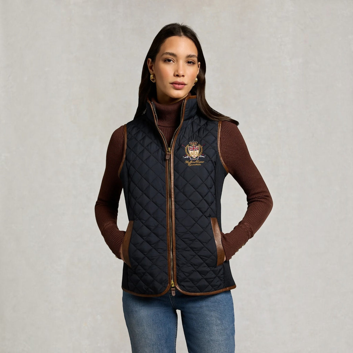 A woman modeling a black quilted zip-front vest with brown trim and a small embroidered crest logo on the left chest, worn over a brown turtleneck and blue jeans against a light gray textured background.