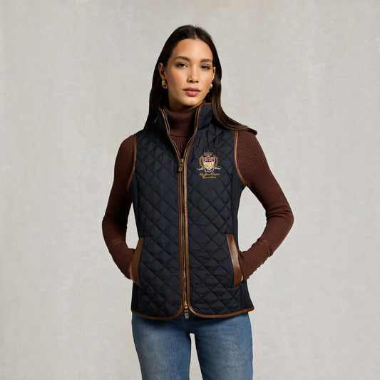 A woman modeling a black quilted zip-front vest with brown trim and a small embroidered crest logo on the left chest, worn over a brown turtleneck and blue jeans against a light gray textured background.