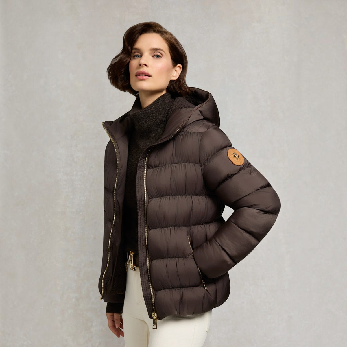 A woman models a dark brown puffer jacket with a high collar, gold-tone zipper, and a circular brown logo patch on the sleeve, set against a textured light gray background.