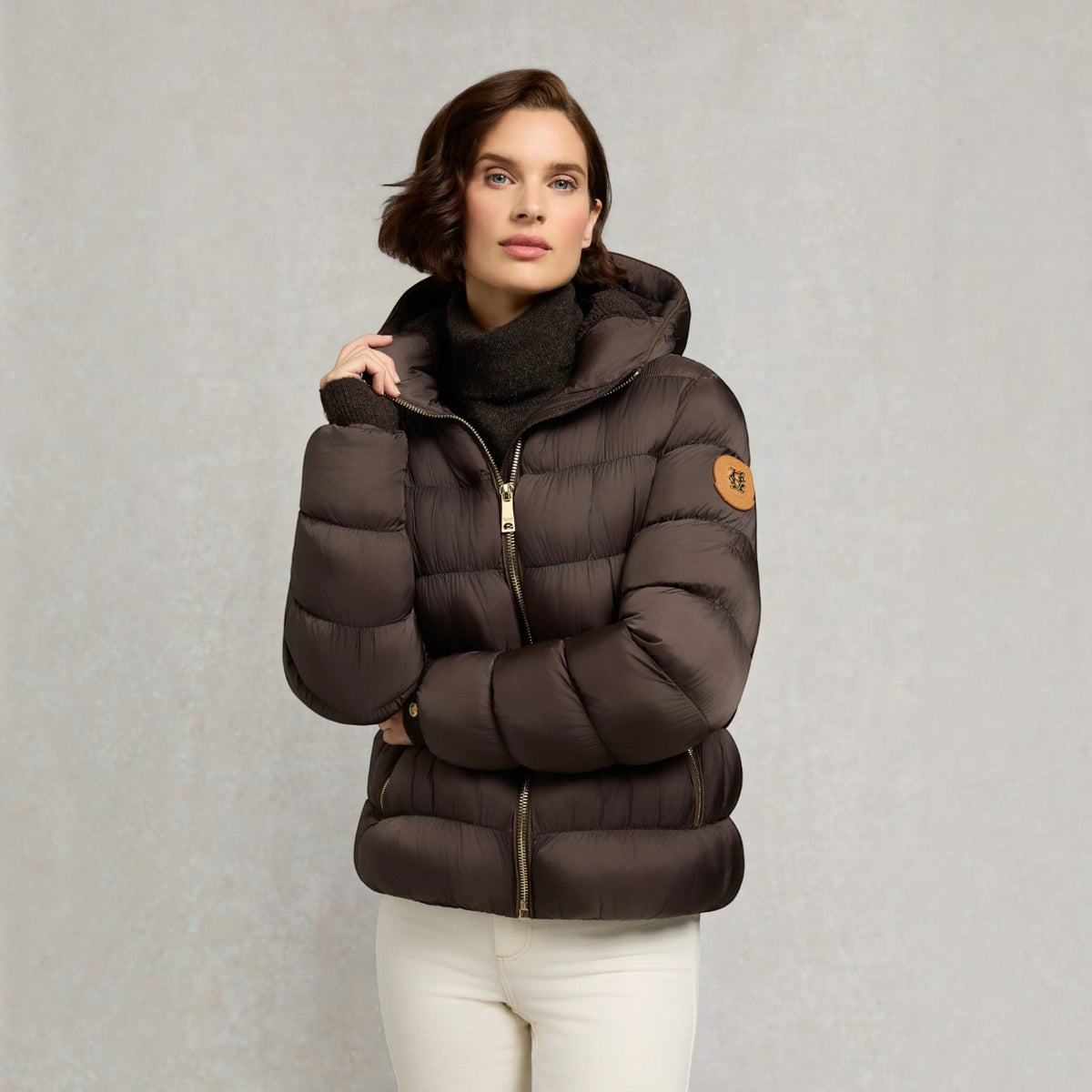 A woman wearing a dark brown puffer jacket with a high collar, gold zipper, and a circular tan patch on the sleeve, paired with a black turtleneck and white pants, set against a textured gray background.