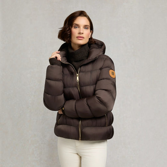 A woman wearing a dark brown puffer jacket with a high collar, gold zipper, and a circular tan patch on the sleeve, paired with a black turtleneck and white pants, set against a textured gray background.