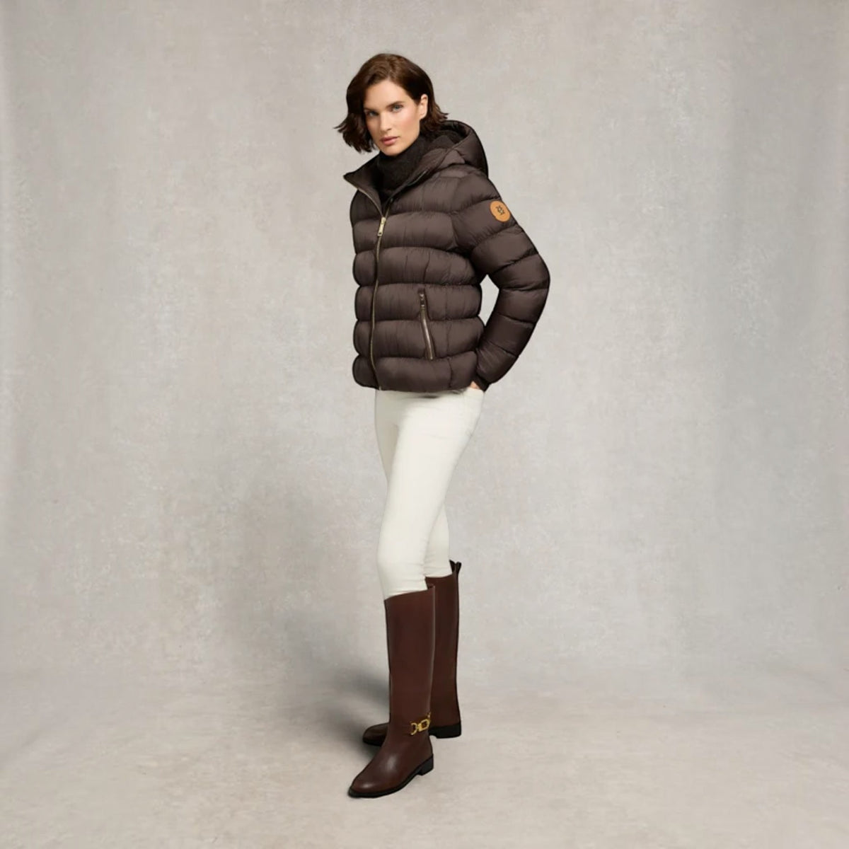 A woman models a dark brown puffer jacket with a small orange logo patch on the sleeve, paired with white pants and brown knee-high boots, against a textured gray studio background.