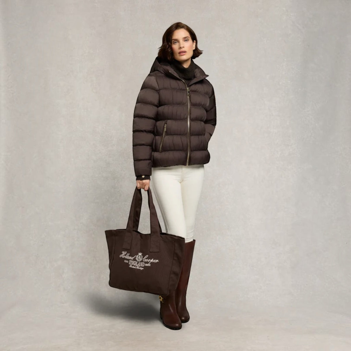 A woman stands against a textured light gray studio backdrop wearing a dark brown puffer jacket, white pants, and brown knee-high boots, while holding a dark brown tote bag with white script text.