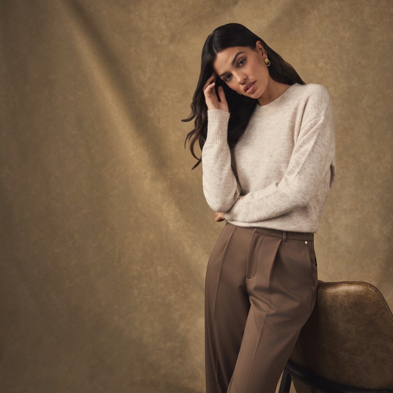 A woman posing in a studio setting, wearing a light cream knit sweater and brown trousers, leaning against a textured brown chair against a matching brown backdrop.