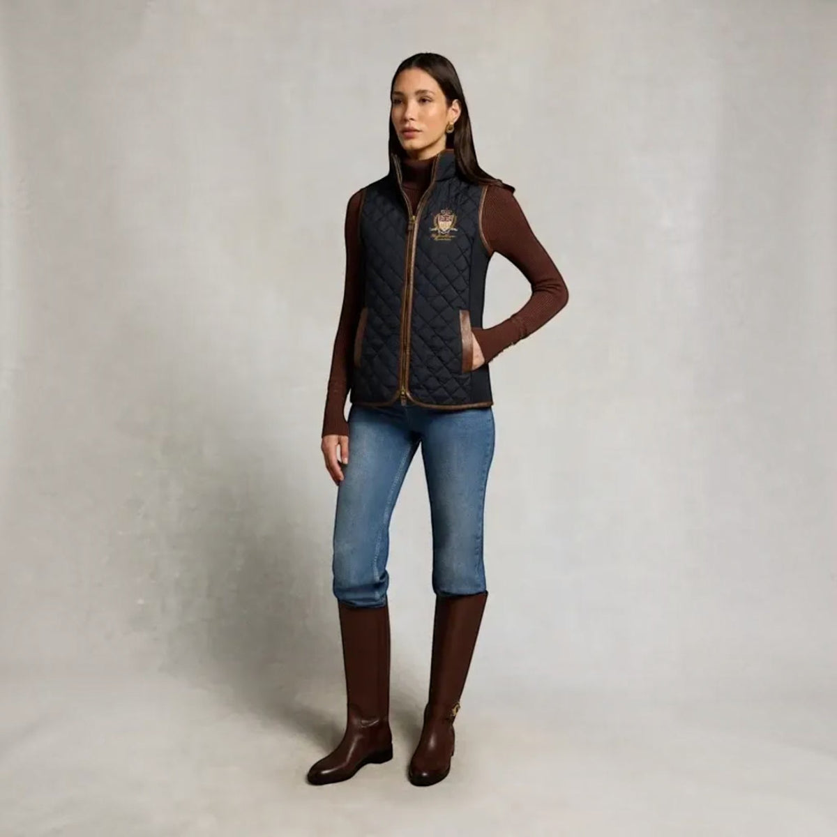 A woman models a dark navy quilted vest with brown trim over a brown long-sleeve top, blue cropped jeans, and brown riding boots, standing against a neutral studio backdrop.