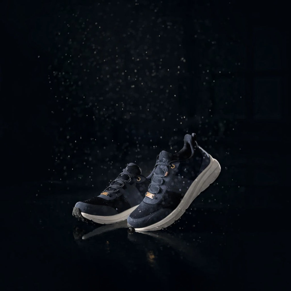 A pair of dark navy and black athletic sneakers with subtle gold accents, shown floating above a reflective dark surface with a dramatic water droplet effect in the background.