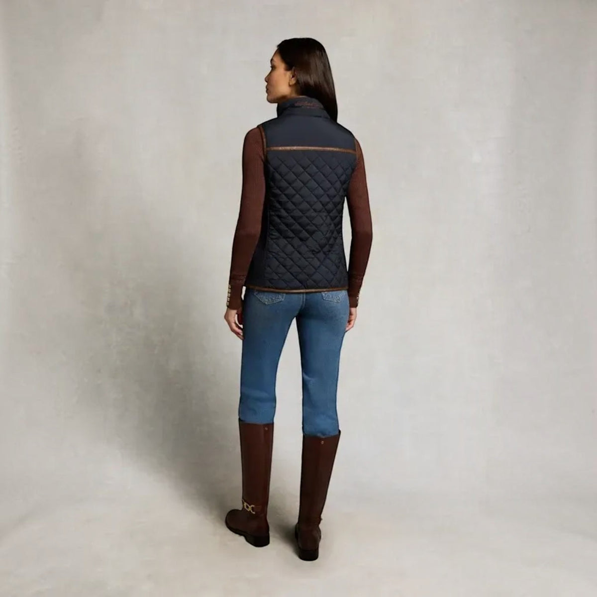 A woman is shown from the back wearing a dark navy quilted vest over a brown long-sleeve top, blue jeans, and brown knee-high boots, standing against a neutral textured studio backdrop.