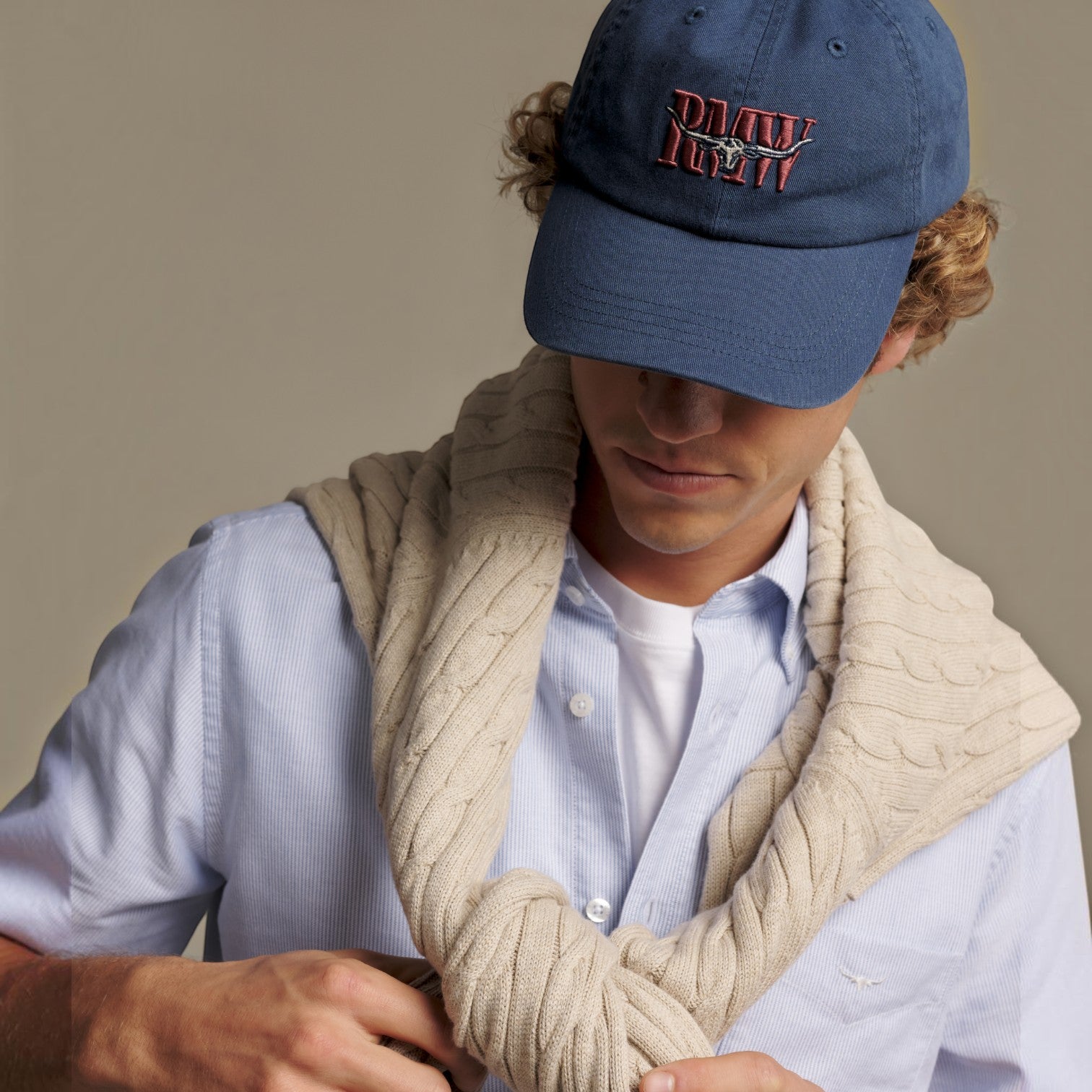 A man wearing a blue baseball cap with 'RMW' embroidered on the front, a light blue button-down shirt, and a thick cream-colored cable-knit scarf, posed against a solid beige background.