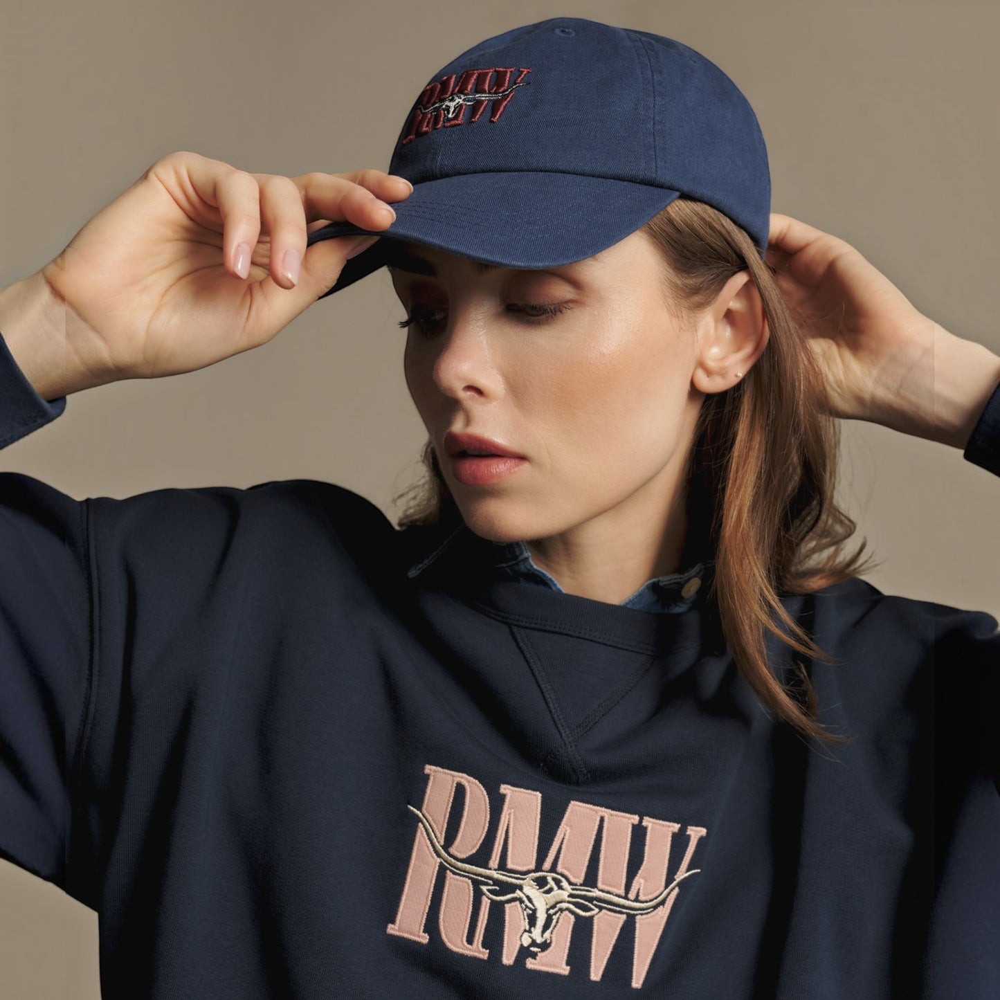A woman wearing a navy blue baseball cap with red embroidered text and a matching navy sweatshirt with a pink graphic, posing against a neutral beige background.