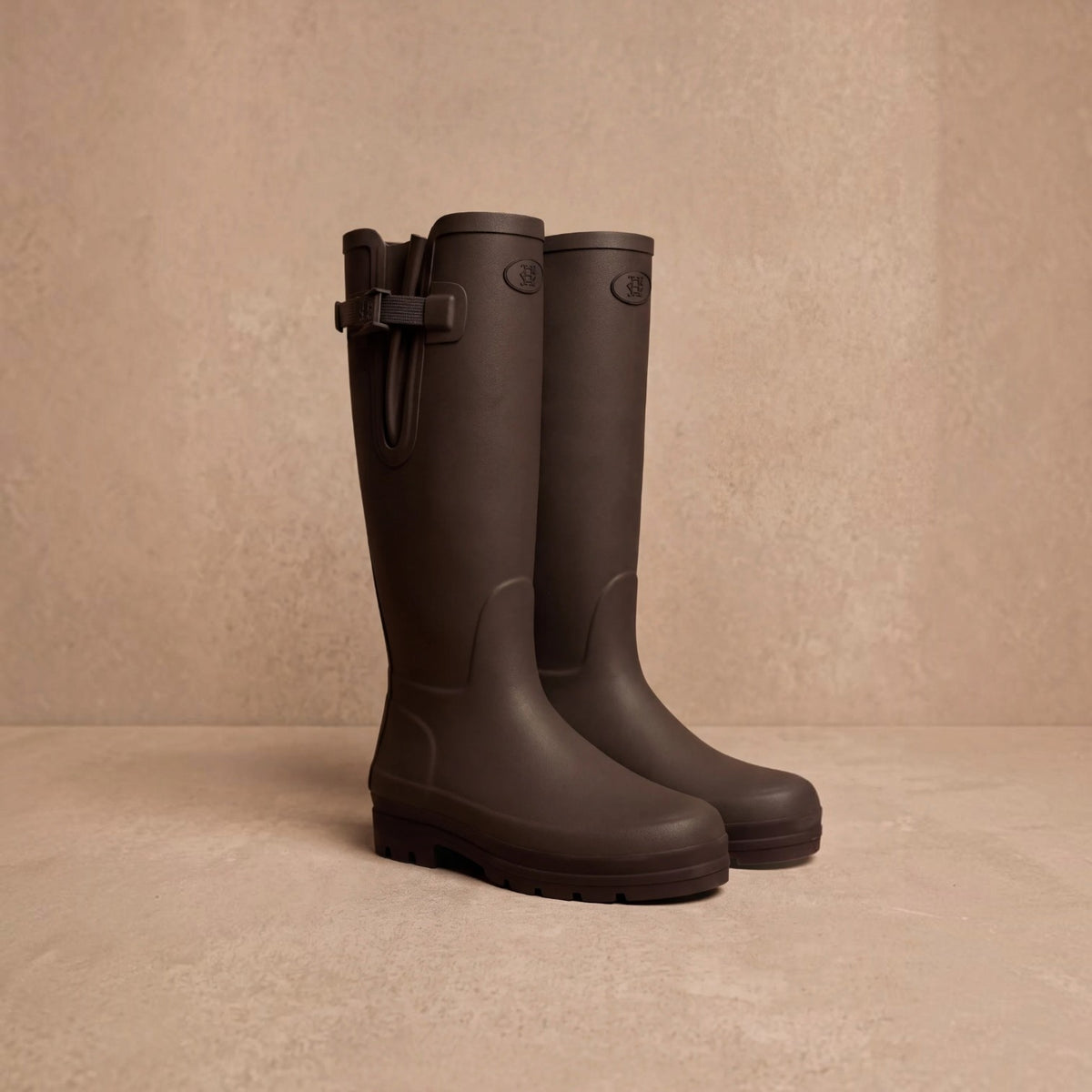 A pair of dark brown rubber rain boots with buckle straps, displayed on a matching beige textured surface against a beige background.