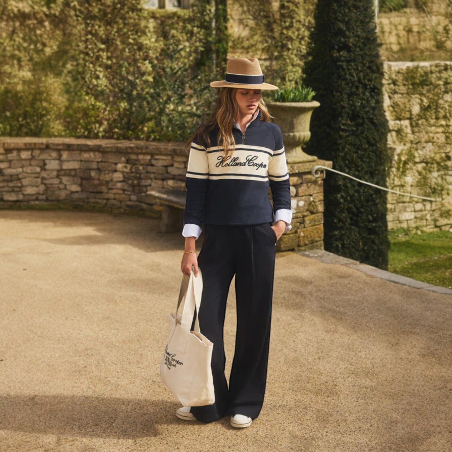 Woman in a garden wearing a navy and white striped sweater with 'Rolland Lenon' branding, holding a tote bag.