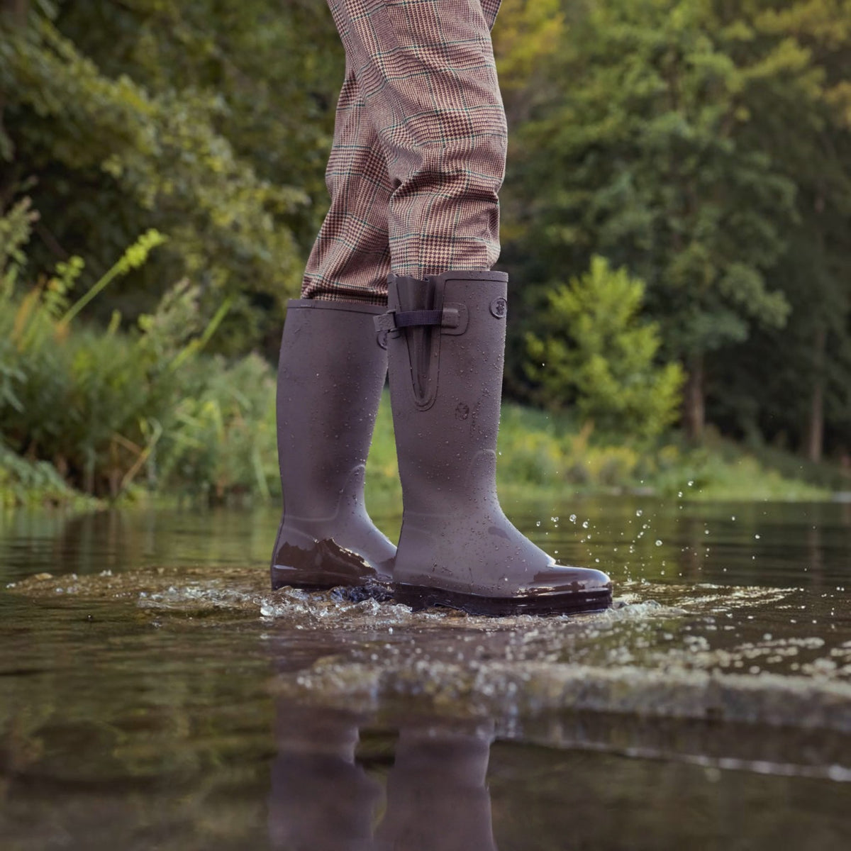 A person wearing dark brown rubber rain boots and brown houndstooth trousers standing in shallow water outdoors, with trees and greenery blurred in the background.