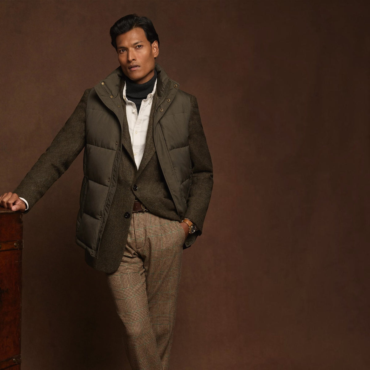 A man models a dark olive green puffer vest over a brown waistcoat and white shirt, paired with brown houndstooth trousers, set against a solid dark brown studio background.