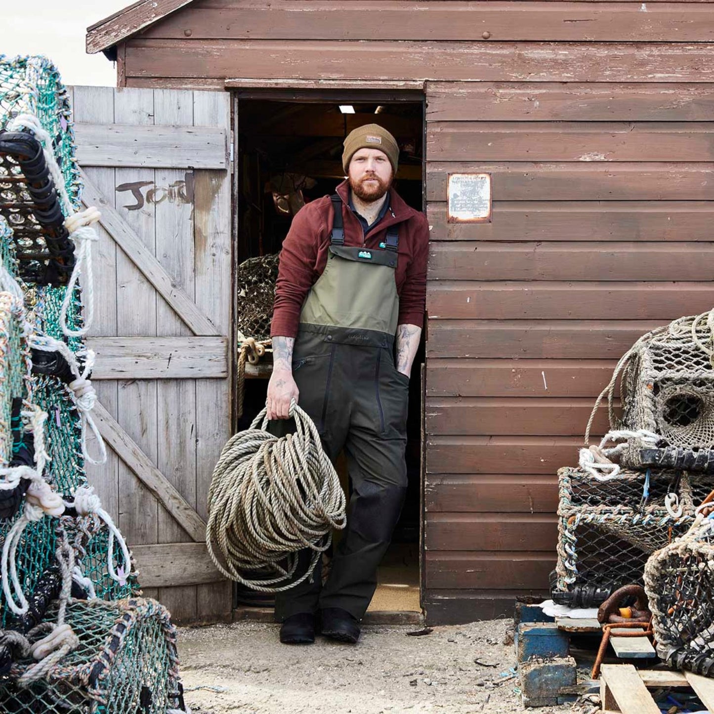 A man wearing a brown beanie, maroon long-sleeve shirt, olive green and black overalls, and black boots stands in the doorway of a rustic brown wooden shed, holding a large coil of rope. Fishing nets and buoys are stacked on both sides of the doorway.
