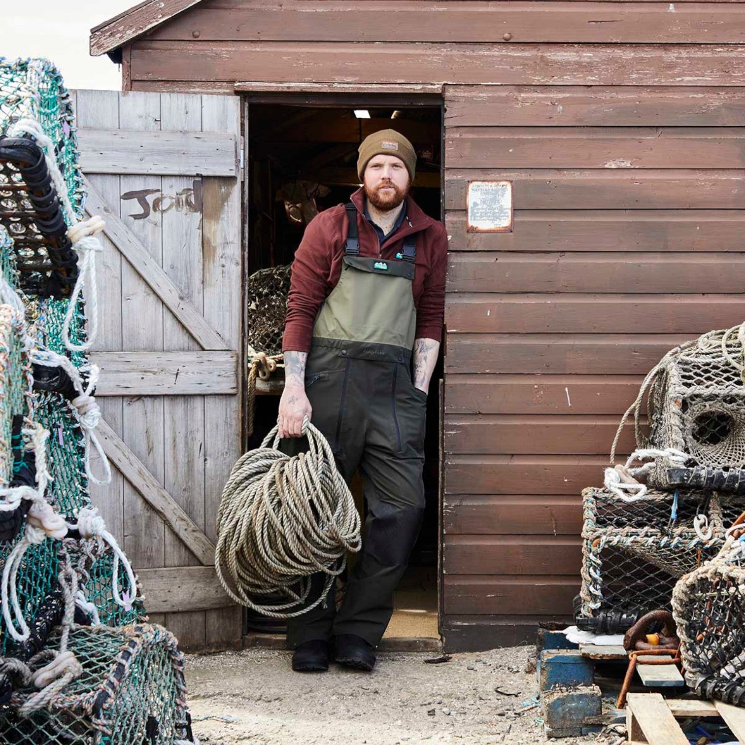 A man wearing a brown beanie, maroon long-sleeve shirt, olive green and black overalls, and black boots stands in the doorway of a rustic brown wooden shed, holding a large coil of rope. Fishing nets and buoys are stacked on both sides of the doorway.