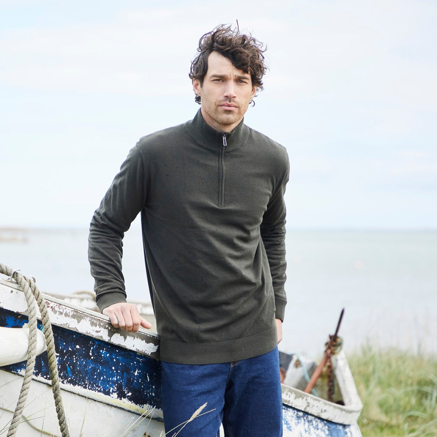 A man wearing an olive green quarter-zip long-sleeve sweater and blue jeans stands next to a weathered blue boat with a body of water and grass in the background.