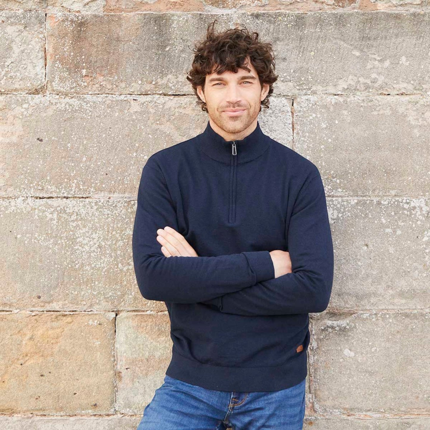 A man with curly brown hair wearing a navy blue quarter-zip pullover sweater and blue jeans, standing with arms crossed in front of a rustic stone wall.