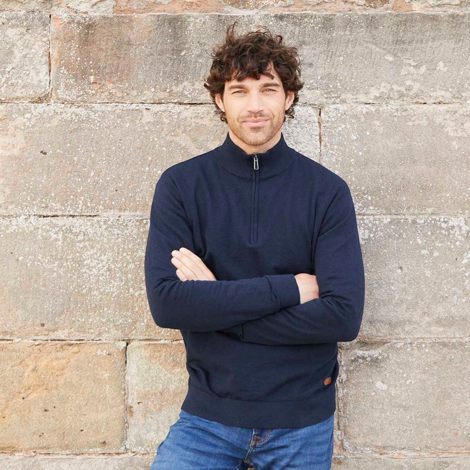 A man with curly brown hair wearing a navy blue quarter-zip pullover sweater and blue jeans, standing with arms crossed in front of a rustic stone wall.