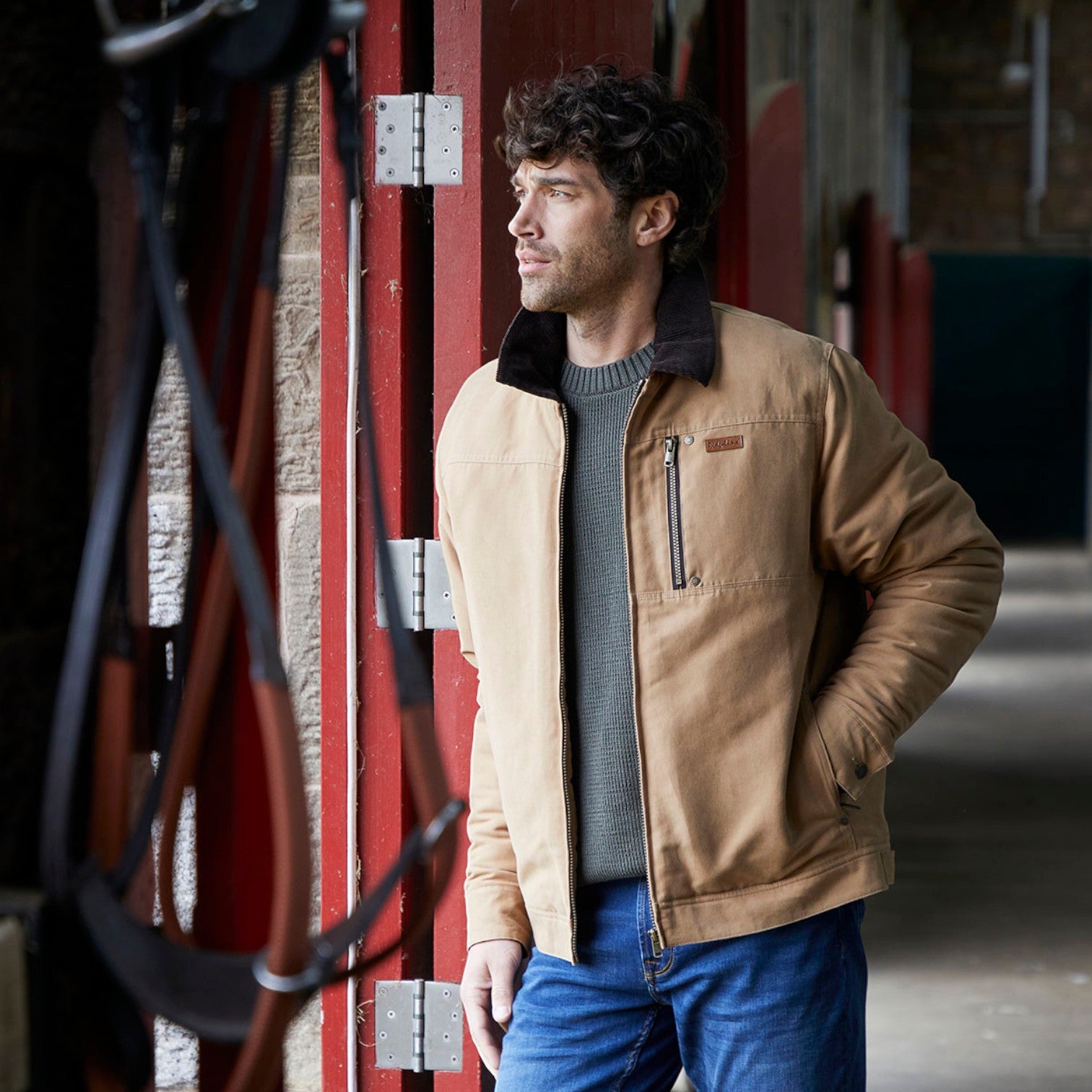 A man wearing a tan canvas utility jacket with a dark brown fleece collar, layered over a gray knit sweater and blue jeans, standing in a rustic corridor with red metal doors.