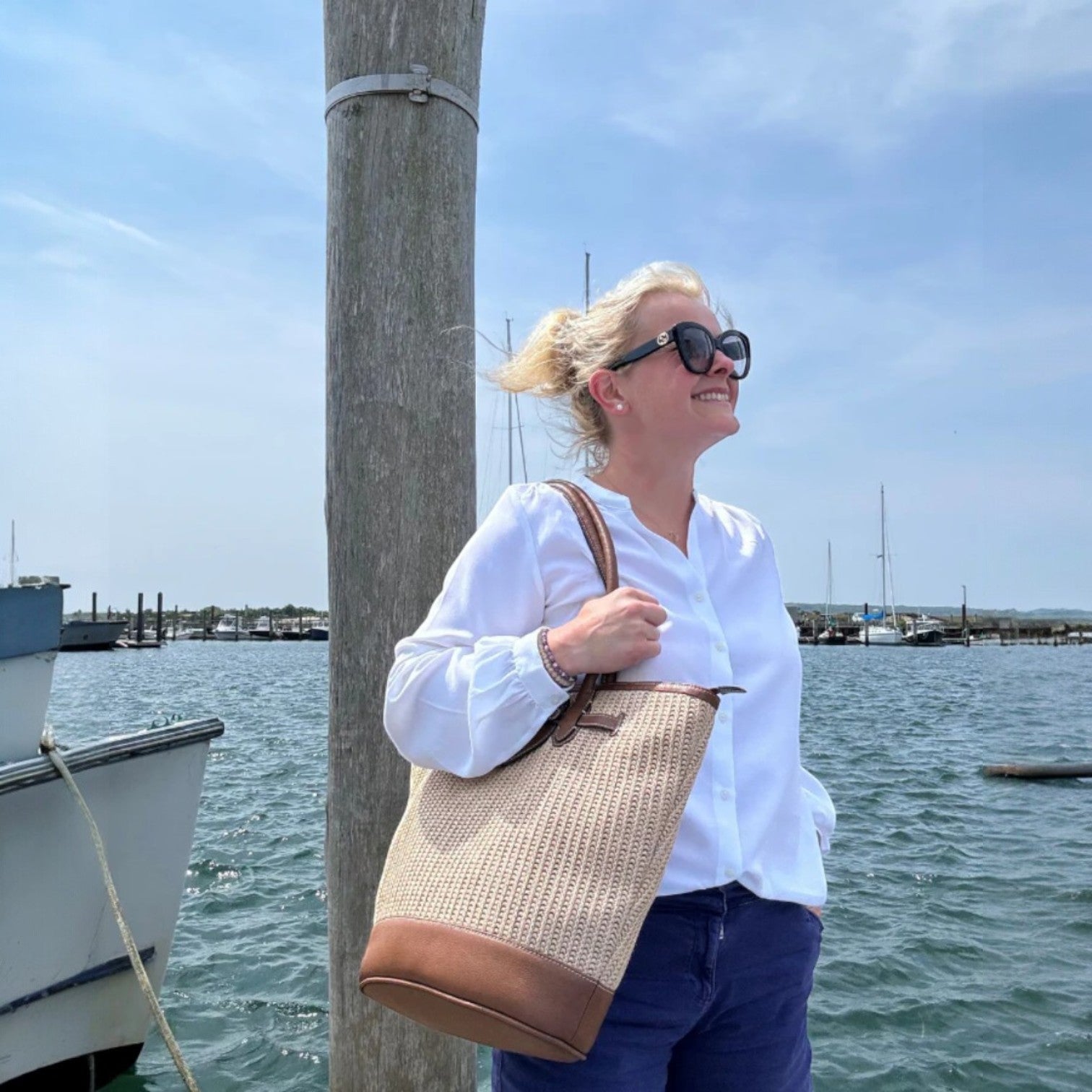 Woman holding a woven bag by a dock with boats and water in the background