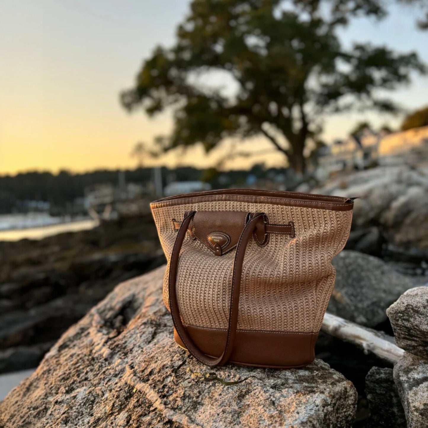 Woven handbag with brown leather accents on a rock by a waterfront at sunset.