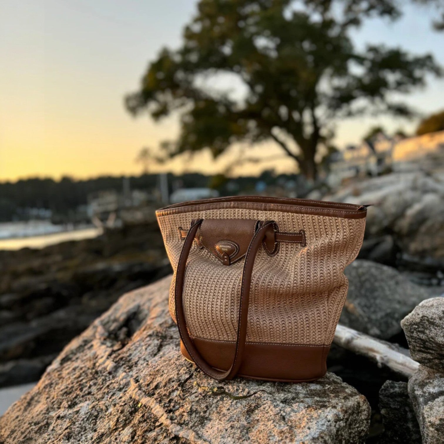 Woven handbag with brown leather accents on a rock by a waterfront at sunset.