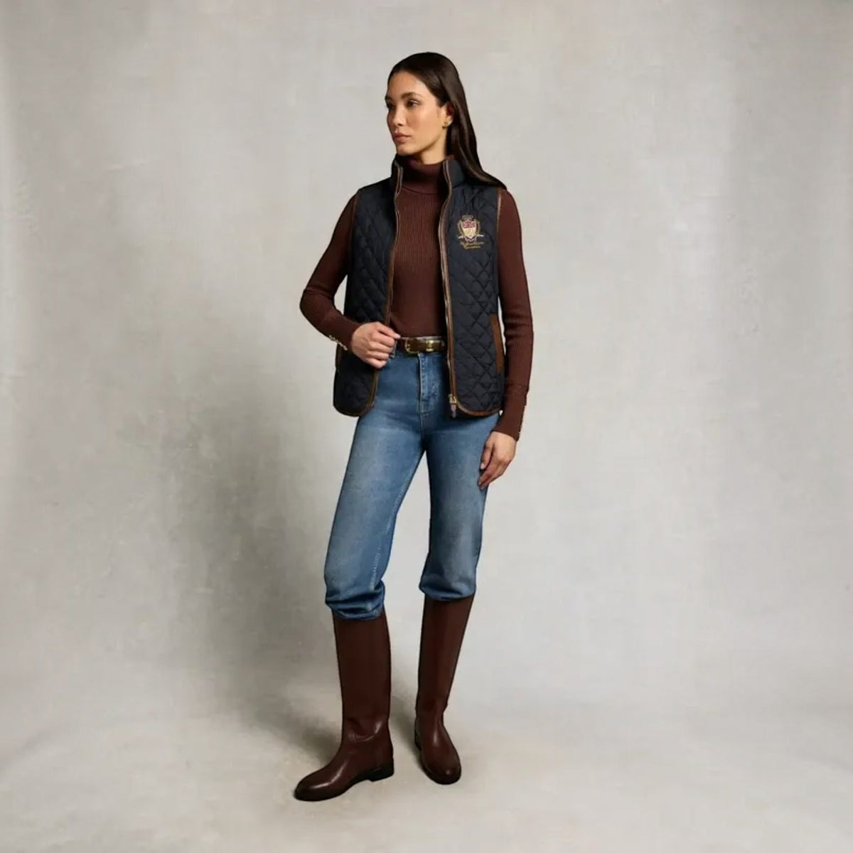 A woman stands against a neutral studio backdrop wearing a quilted dark navy vest over a brown turtleneck, blue jeans, and brown knee-high boots. A small embroidered crest is visible on the left chest of the vest.